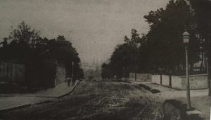 view of muddy road, lined by garden walls and trees, sloping down hill.